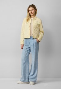 Geel denimjasje met knoopsluiting en dubbele borstzakken, gecombineerd met lichtblauwe wijde jeans en witte sneakers. Gladde textuur.
