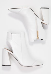 Botas de tobillo de cuero blanco con tacón, punta afilada, laterales abiertos y cremallera en la parte trasera. Los tacones son de estilo bloque con un acento metálico.