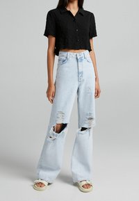 Persona che indossa jeans larghi di un blu chiaro strappati, una maglietta nera a maniche corte e tagliata, e sandali beige, in piedi davanti a uno sfondo grigio uniforme.