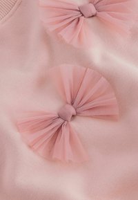 Material din bumbac roz, cu două funde din tul roz, având noduri centrale. Textură moale cu un finisaj neted de tricot, evidențiind detalii delicate.