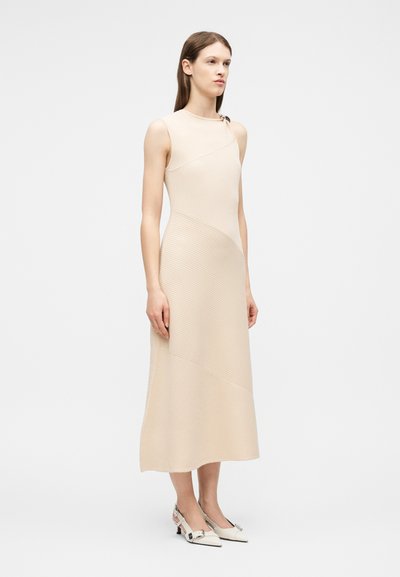 Femme debout portant une robe midi sans manches beige côtelée avec des coutures asymétriques et des chaussures blanches à talons bas pointues avec des boucles.