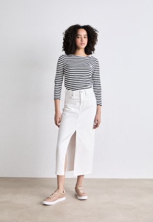 Kvinde med krøllet hår iført en sort og hvid stribet top, hvid midi nederdel med slids foran og beige sneakers, stående foran en enkel baggrund.