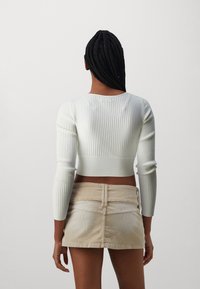 Žebrovaný bílý krátký svetr s dlouhými rukávy, spárovaný s béžovou manšestrovou mini sukní. Materiál vypadá měkce a má přiléhavý design.