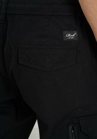 Schwarze Cargo-Hosen aus strukturiertem Stoff, mit einer Gesäßtasche mit Klappe und einem kleinen Markenlabel, das auf der Tasche aufgenäht ist.