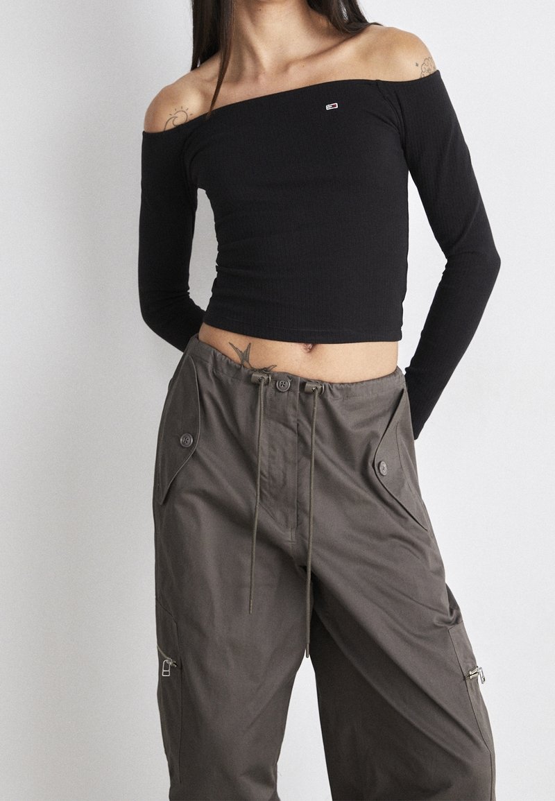 Schwarzes, geripptes, schulterfreies Langarmoberteil kombiniert mit olivgrünen Cargo-Hosen, die über Knopftaschen und einen Kordelzug an der Taille verfügen.