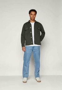 Giacca bottonata verde scuro con due tasche sul petto, indossata sopra una maglietta bianca. Abbinata a jeans azzurri chiari e sneakers bianche con suola beige.