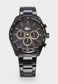 Montre en métal noir avec un cadran rond, des accents dorés, trois sous-cadrans et une lunette tachymétrique. Elle dispose d'un bracelet à maillons et du logo Lacoste.
