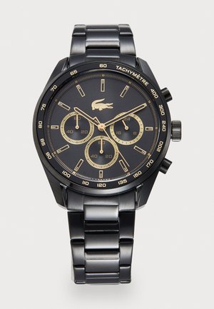 Montre en métal noir avec un cadran rond, des accents dorés, trois sous-cadrans et une lunette tachymétrique. Elle dispose d'un bracelet à maillons et du logo Lacoste.