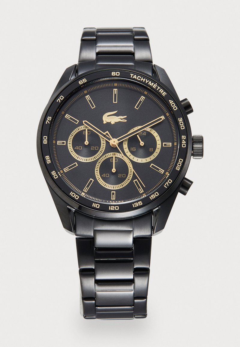 Montre en métal noir avec un cadran rond, des accents dorés, trois sous-cadrans et une lunette tachymétrique. Elle dispose d'un bracelet à maillons et du logo Lacoste.