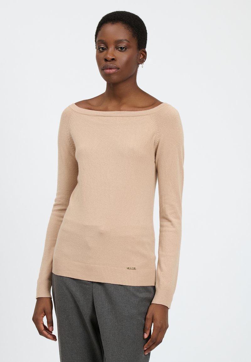 Maglione beige a maniche lunghe con scollo a barca, orlo a costine e una leggera lucentezza. Presenta un piccolo accento del logo in metallo vicino al fondo.