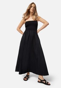 Abito maxi nero con busto smock e spalline sottili; il tessuto fluido scende fino alla caviglia. Indossato con sandali neri con cinturini.