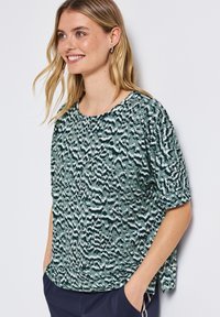 Nuori nainen hymyilee, pukeutuneena vihreä-mustaraidalliseen lyhythihaiseen puseroon ja tummansinisiin housuihin, seisten kädet taskuissa valkoista taustaa vasten.