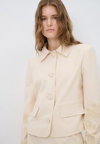 Beige jas met een gestructureerd ontwerp, voorzien van een kraag, zeven knopen, twee voorzakken en geborduurde bloemaccenten op de mouwen.