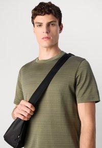 Mlad moški v olivno obarvani srajci s kratkimi rokavi drži črn trak torbice čez ramo pred enotno ozadjem.