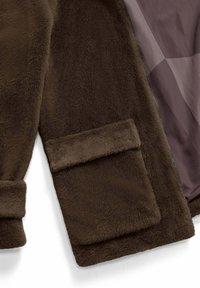Bruine faux fur jas met een zachte textuur, voorzien van een rits en een klepzak. De voering aan de binnenkant is een lichtere tint bruin.