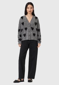 Cardigan grigio a maglia con motivi a cuore neri, maniche lunghe, scollatura a V profonda e chiusura anteriore con bottoni. Abbinato a jeans neri a vita alta.