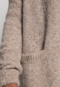 Pull beige en maille avec une texture douce, présentant une poche visible et des poignets côtelés. Le tissu semble chaud et confortable.