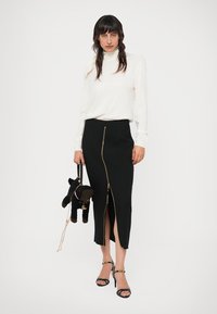 Mustat vetoketjuhameet, joissa on korkea halkio, yhdistettynä pitkähiharaiseen valkoiseen toppiin. Malli pitää pientä pehmeää koirakassia käsissään. Avoimet sandaalit korkokengillä.