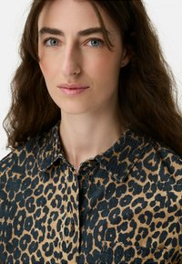 Frau mit blauen Augen und langen braunen Haaren, die ein geknöpftes Hemd mit Leopardenmuster und Kragen vor einem hellen Hintergrund trägt.
