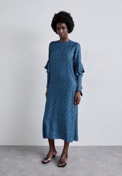 Langes blaues Kleid mit Blumenmuster, Rüschensäumen und geradem Schnitt. Trägt dazu schwarze Sandaletten mit Riemen. Glatter Stofftextur.