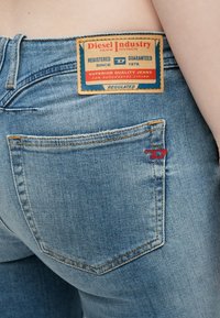 Calças de ganga azul com uma etiqueta de pele castanha com texto em vermelho e azul nas costas. Bolso com costura dupla e um destaque de logótipo vermelho.