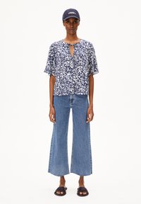 Marineblaues, kurzärmliges Oberteil mit floralen Mustern und einem Schlüsselloch-Ausschnitt, kombiniert mit hochtaillierten, weiten blauen Jeans und dunklen Sandalen.