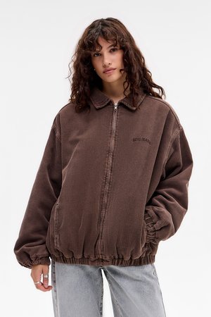 Vrouw met krullend haar, gekleed in een bruine corduroy ritsjack en lichtblauwe jeans, staand met handen in de zakken tegen een witte achtergrond.