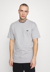 Camiseta de algodón gris con mangas cortas, escote redondo y un pequeño logo negro de Vans en el pecho. Textura suave y corte relajado.