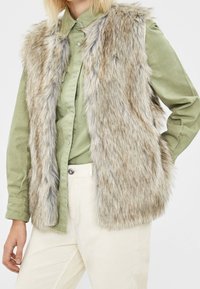 Gilet en fausse fourrure dans des tons beige et gris, avec un col montant et une ouverture sur le devant, porté sur une chemise verte à boutons et un pantalon couleur crème.