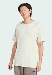 T-shirt beige clair avec des manches raglan courtes, affichant un petit logo embossé sur la poitrine et une coupe décontractée. Texture douce.