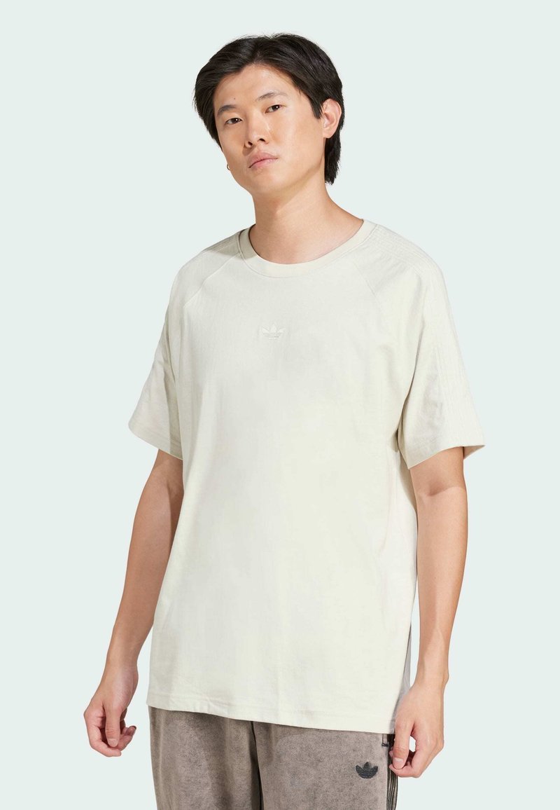 T-shirt bege claro com mangas raglan curtas, apresentando um pequeno logótipo em relevo no peito e um ajuste descontraído. Textura suave.