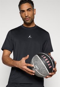 Μαύρο αθλητικό t-shirt με κοντά μανίκια και λευκό λογότυπο Jordan, κρατημένο από άτομο που κρατάει μια ασπρόμαυρη μπασκέτα με σχέδια.