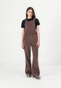 Salopette di velluto a coste marrone con design a gamba dritta, spalline regolabili e tasche laterali, indossata sopra una maglietta nera.