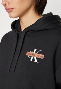 Persoon draagt een zwarte hoodie met het Calvin Klein Jeans-logo op de borst en trekkoorden zichtbaar bij de halslijn.