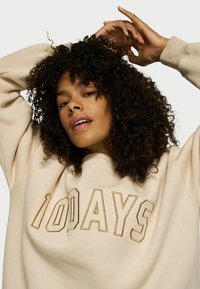 Sudadera beige con "100 DÍAS" bordado en dorado. Presenta un ajuste holgado y una tela texturizada. El cabello rizado enmarca parcialmente la cara.