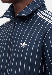 Primo piano di una persona che indossa una giacca blu navy con zip, con sottili righe bianche, tre strisce bianche sulle spalle e un piccolo logo Adidas bianco sul petto.