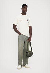 Man wearing white graphic t-shirt, loose faded green jeans, white sneakers, holding green crossbody bag by strap.