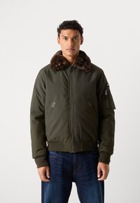 Olivengrøn bomberjakke med faux fur-krave, lynlås lukning og sidelommer. Inkluderer detaljer på den ene ærme. Bæres med mørke jeans.