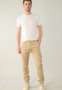 Beige cargobroek met elastische boorden, meerdere zijzakken en een gladde textuur. Gecombineerd met een eenvoudig wit T-shirt, staande op een lichte achtergrond.