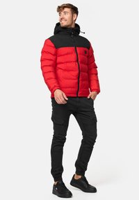 Rød og sort polstret jakke med lynlås, hætte og elastiske manchetter. Kombineret med sorte cargo bukser og sorte sneakers.