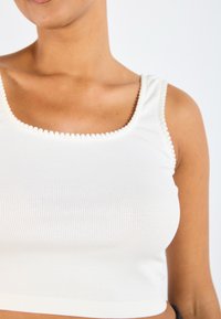 Hvit ribbestrikka crop-top med firkantet hals, med teksturert bølget kant langs kantene. Stoffer ser mykt og tettsittende ut.