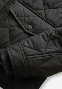 Black quilted jacket featuring a button closure and ribbed cuff. The fabric has a smooth texture with diamond-shaped stitching accents.