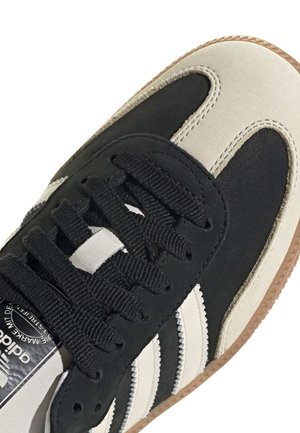Zbliżenie czarno-beżowego buta sportowego Adidas z czarnymi sznurowadłami i gumową podeszwą, pokazującego trzy białe charakterystyczne paski z boku.