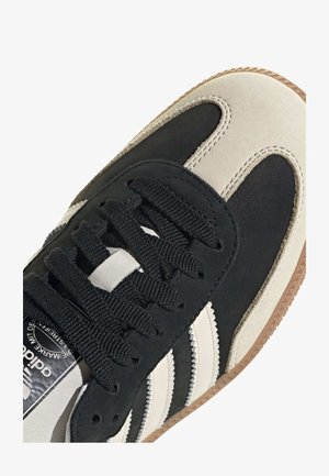 Zbliżenie czarno-beżowego buta sportowego Adidas z czarnymi sznurowadłami i gumową podeszwą, pokazującego trzy białe charakterystyczne paski z boku.