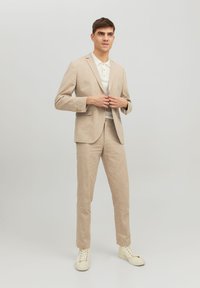 Completo in lino beige con blazer aderente e pantaloni affusolati, abbinato a una polo bianca e scarpe da ginnastica chiare, su uno sfondo neutro.