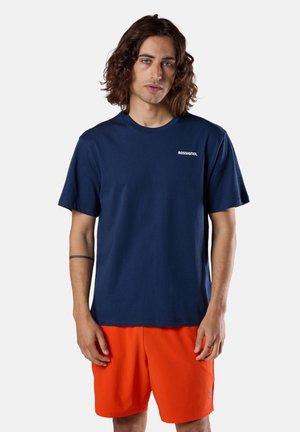 Mężczyzna o włosach do ramion, ubrany w granatową koszulkę Rossignol i jaskrawo pomarańczowe spodenki, stojący na tle jednolitego, jasnego tła.