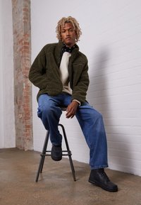 Giacca in felpa verde oliva con tasca con zip, maglione crema sotto, jeans blu e sneakers nere su uno sgabello di metallo contro un muro bianco.