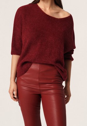 Kobieta ubrana w luźny bordowy sweter z opuszczonymi ramionami oraz dopasowane, wysokiej talii bordowe skórzane spodnie, stojąca na jednolitym tle.