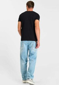 Homme de dos portant un t-shirt noir, un jean bleu clair et des baskets grises, sur fond uni.