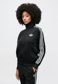 Must sportlik jakk kõrge kraega, valgete kolmekordsete triipudega varrukatel ja väike valge logo vasakul rinnal. Sile tekstuur.